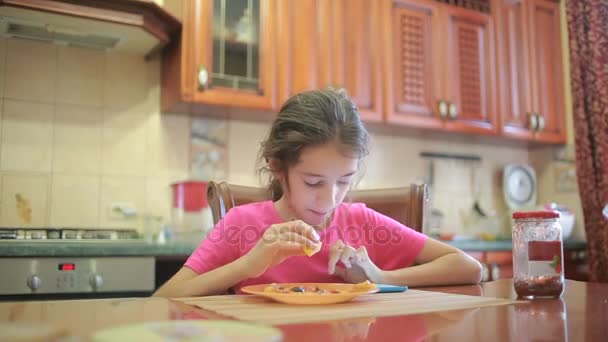 Дівчина їсть піцу та фрукти, граючи з телефоном на кухні за столом — стокове відео