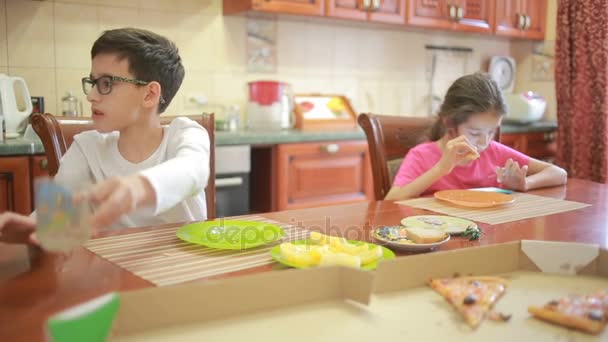 Niño y niña, gemelos comen pizza y fruta mientras juegan con el teléfono en la cocina en la mesa — Vídeos de Stock