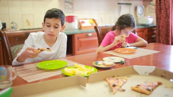 Menino e menina, gêmeos comem pizza e frutas enquanto brincam com telefone na cozinha na mesa — Vídeo de Stock