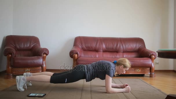 Une femme au foyer fait des exercices de fitness pour la première fois, en regardant la tablette et la télévision, c'est difficile et inconnu, elle est fatiguée . — Video