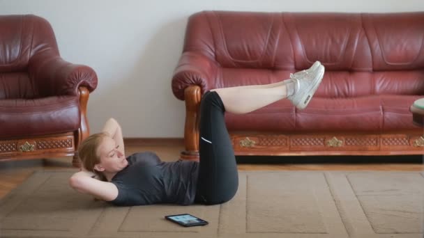 Une femme au foyer fait des exercices de fitness pour la première fois, en regardant la tablette et la télévision, c'est difficile et inconnu, elle est fatiguée . — Video