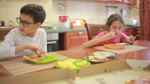 Menino e menina, gêmeos comem pizza e frutas enquanto brincam com telefone na cozinha na mesa — Vídeo de Stock