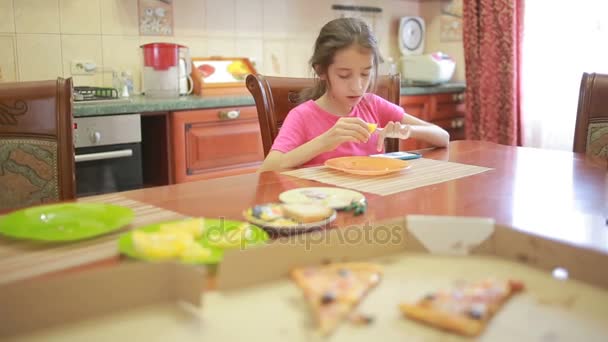 Menina comendo pizza e frutas brincando com telefone na cozinha à mesa — Vídeo de Stock