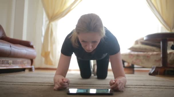 Eine Hausfrau macht zum ersten Mal Fitnessübungen, schaut auf Tablet und Fernseher, es ist schwierig und ungewohnt, sie ist müde. — Stockvideo