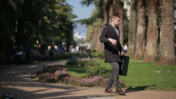 Šťastný podnikatel v kabátě a s kufrem tančí šťastně venku na pozadí palem. — Stock video