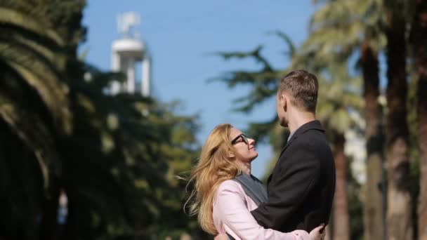 Pareja feliz enamorada divirtiéndose. Linda mujer hermosa con un hombre guapo en un abrigo besándose. Sobre el telón de fondo de palmeras y árboles . — Vídeos de Stock