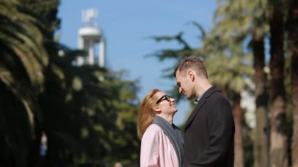 Coppia felice innamorata divertirsi. Bella donna carina con un bell'uomo in un cappotto che si bacia. Sullo sfondo di palme e alberi . — Video Stock