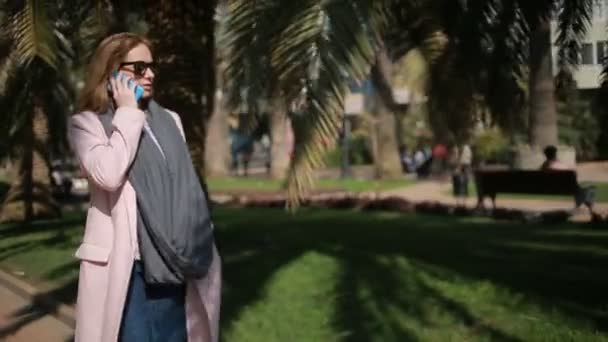 Молодая женщина улыбается, разговаривает по мобильному телефону в парке, прогуливаясь по аллее с пальмами — стоковое видео