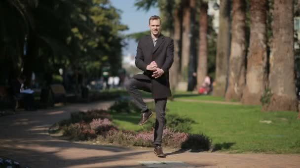 Happy businessman in a coat and with a suitcase dancing happily outdoors against a backdrop of palm trees. — Stock Video