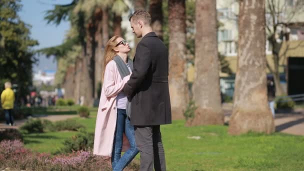 Coppia felice innamorata divertirsi. Bella donna carina con un bell'uomo in un cappotto che si bacia. Sullo sfondo di palme e alberi . — Video Stock