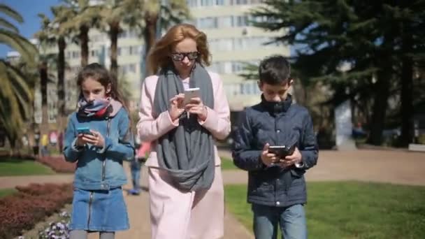 Mulher com filhos. Mãe e crianças estão andando ao longo do beco, todo mundo está ocupado com seu telefone — Vídeo de Stock