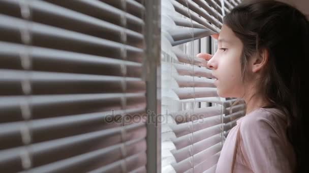 Niña mira a la calle desde la ventana a través de las persianas — Vídeos de Stock