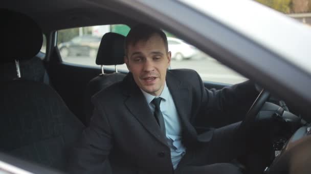 Un hombre de negocios se sienta al volante de un coche y habla con alguien en la calle, invitando con él. Sonriente, amigable — Vídeos de Stock