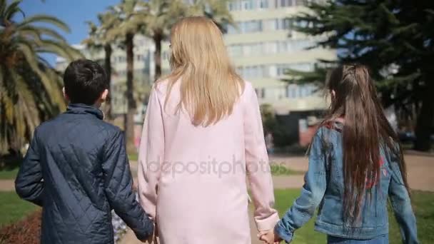 Una donna con figli, una madre che tiene due gemelli per mano passeggiando lungo un viale di palme — Video Stock