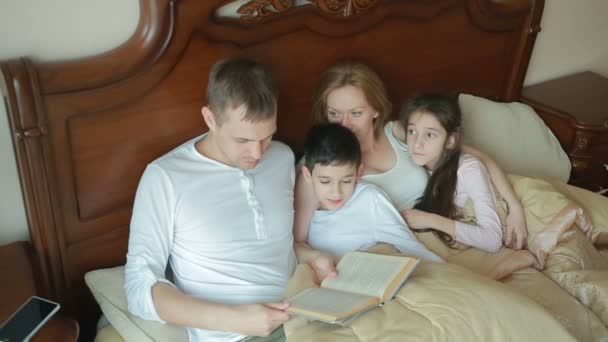Счастливая семья на кровати читает книгу вслух дома — стоковое видео