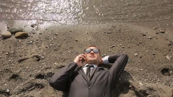 Бизнесмен в костюме и солнечных очках лежит на пляже рядом с линией для серфинга — стоковое видео