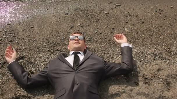 Un hombre de negocios con traje y gafas de sol se encuentra en la playa cerca de la línea de surf — Vídeo de stock
