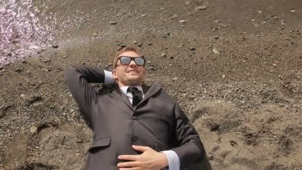 Un uomo d'affari in completo e occhiali da sole si trova sulla spiaggia vicino alla linea di surf — Video Stock