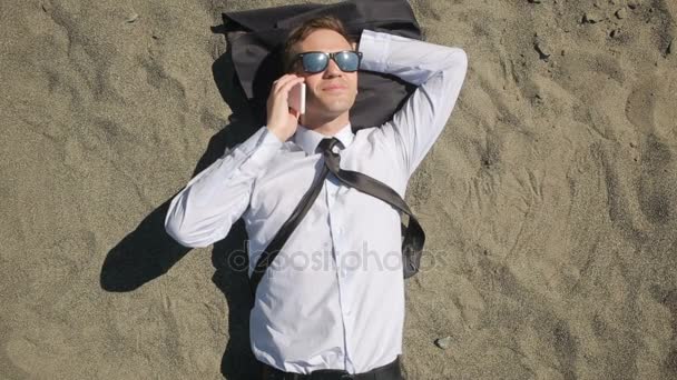 Un hombre de negocios con traje y gafas de sol se encuentra en la playa cerca de la línea de surf — Vídeo de stock