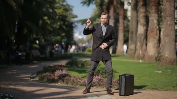 Glad affärsman i en kappa och med en resväska som dansar glatt utomhus mot en bakgrund av palmer. — Stockvideo