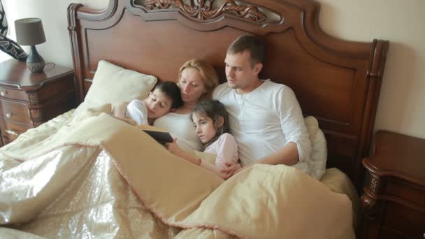 Счастливая семья на кровати читает книгу вслух дома — стоковое видео