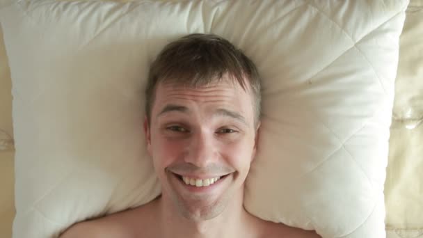Liegt der Mann auf dem Bett und öffnet scharf die Augen, blickt in die Kamera und lächelt. Nahaufnahme. Blick von oben — Stockvideo