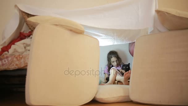 Ein kleines Mädchen sitzt in einem provisorischen Haus aus Kissen und einer Decke — Stockvideo