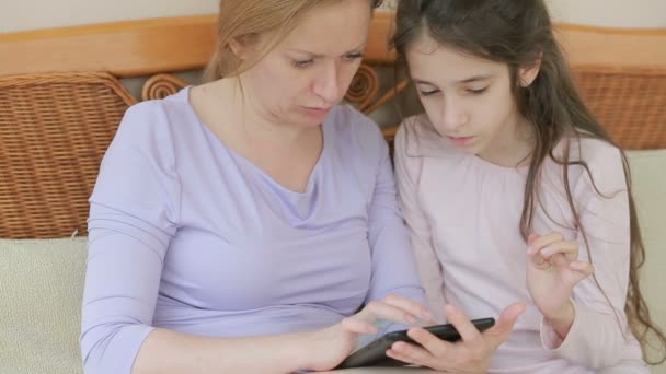 Una hermosa madre y su pequeña hija hablan bien, la madre sostiene una tableta y enseña a su hija — Vídeos de Stock