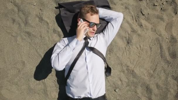 Een zakenman in een pak en zonnebril ligt aan het strand in de buurt van de surf-lijn — Stockvideo