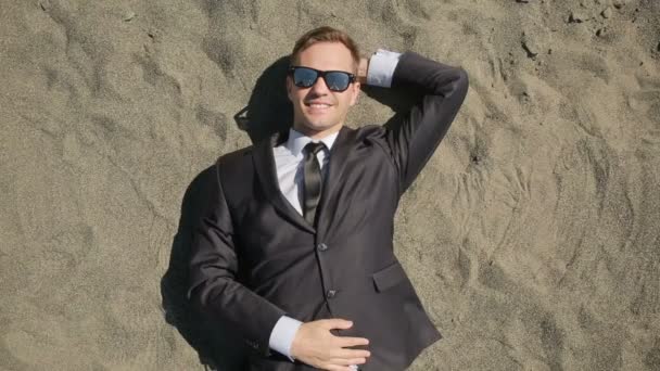 A businessman in a suit and sunglasses lies on the beach near the surf line — Stock Video
