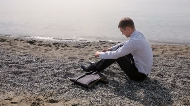 Επιχειρηματίας στο κοστούμι που κάθονται στην παραλία ρίχνει πέτρες στο νερό — Αρχείο Βίντεο