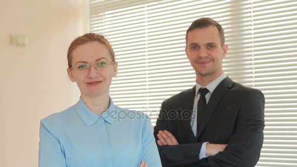 Two businesspeople man and woman are standing and smiling looking into the camera with approval in the office on a window background with shutters. — Stock Video