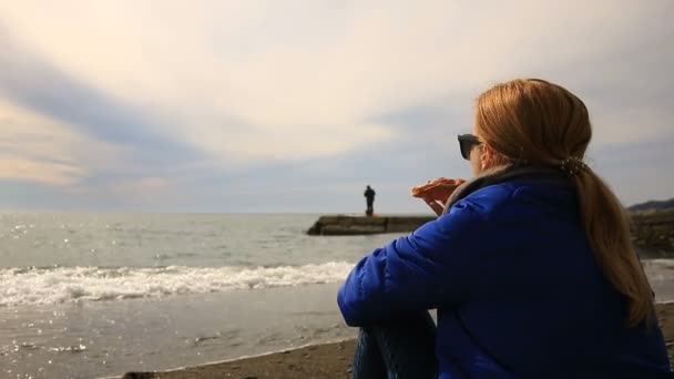 Eine Frau sitzt bei schlechtem Wetter allein an einem einsamen Strand, blickt aufs Meer und isst Pizza — Stockvideo