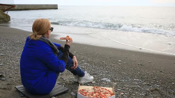 Egy nő egyedül ül egy kihalt strand a rossz időjárás, a tengerre néz, és pizzát eszik — Stock videók