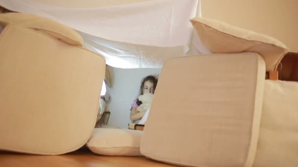 Una niña está sentada en una casa improvisada de almohadas y una casa de mantas — Vídeos de Stock