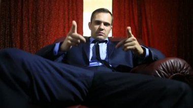 Acımasız adam ile bir deri koltukta oturan, gesticulating ve kameraya bakarak bir takım elbise. Puan parmak bir atış taklit kamera için