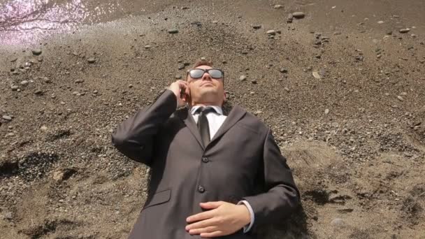 A businessman in a suit and sunglasses lies on the beach near the surf line — Stock Video