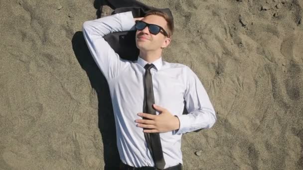 A businessman in a suit and sunglasses lies on the beach near the surf line — Stock Video
