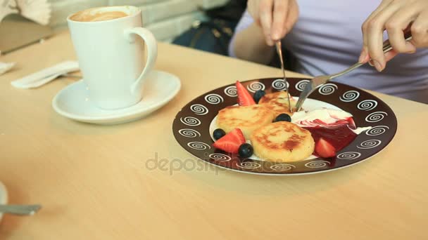 Una chica en un café come panqueques de queso con mermelada de crema agria y bayas frescas con un cuchillo y tenedor — Vídeo de stock