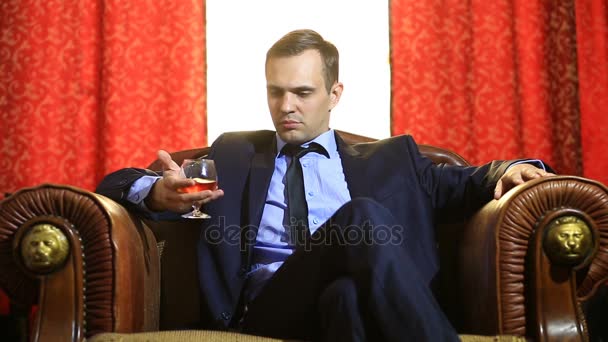 Un uomo brutale in giacca e cravatta si siede su una sedia di pelle con un bicchiere di alcol, guarda la telecamera — Video Stock