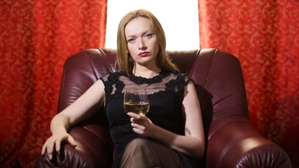 Uma mulher fatal em um vestido preto e um batom vermelho em seus lábios se senta em uma poltrona de couro com um copo de vinho branco e olha arrogantemente para a câmera. Gestos, linguagem corporal — Vídeo de Stock
