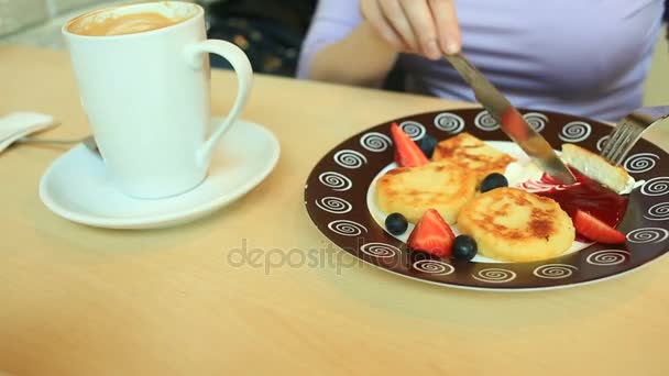 Ein Mädchen in einem Café isst Käsepfannkuchen mit saurer Sahnemarmelade und frischen Beeren mit Messer und Gabel — Stockvideo