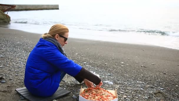 Žena sedí o samotě na opuštěné pláži za špatného počasí, se dívá na moře a žere pizzu — Stock video