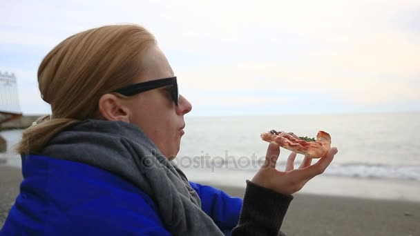 En kvinna sitter ensam på en öde strand i dåligt väder, ser på havet och äter pizza — Stockvideo