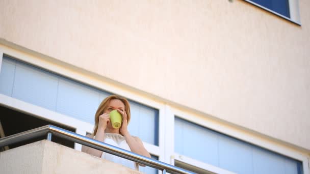 En ung kvinna kliver ut på balkongen och ser sig omkring. Hon ler och stirrar in i distansera. Innehar en mugg med varm dryck — Stockvideo