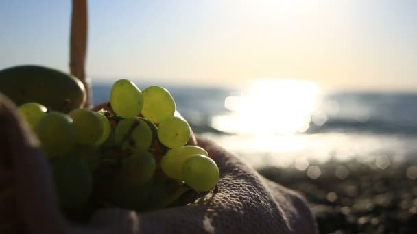 Obstkorb am Strand. die Hand nimmt die Trauben von der Traube vor dem Hintergrund der Meereswellen — Stockvideo