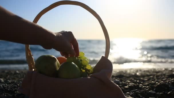 Fruitmand op het strand. De hand neemt de druiven uit de tros druiven tegen de achtergrond van de golven van de zee — Stockvideo