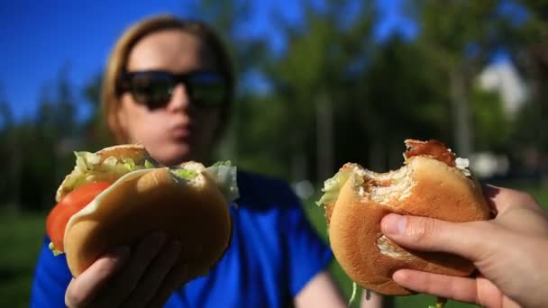 Muž a dívka jíst fast food na trávníku. Oni se ukousnout hamburger od sebe navzájem. Na pozadí rozmazané městské ulice — Stock video