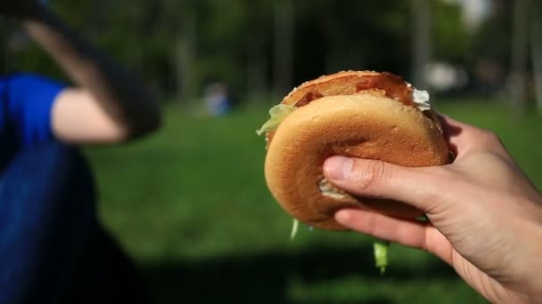 Un hombre y una chica comen comida rápida en el césped. Se muerden la hamburguesa unos a otros. En el contexto de una calle borrosa de la ciudad — Vídeo de stock