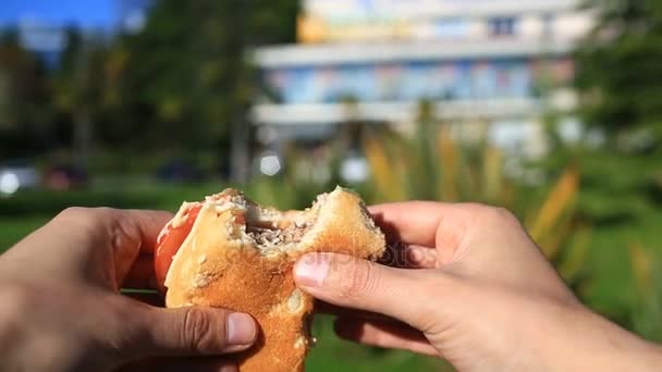 Bir adam sokakta fast food yiyor. Bir hamburger giyer ve yiyor. Bulanık şehir sokak arka planı — Stok video